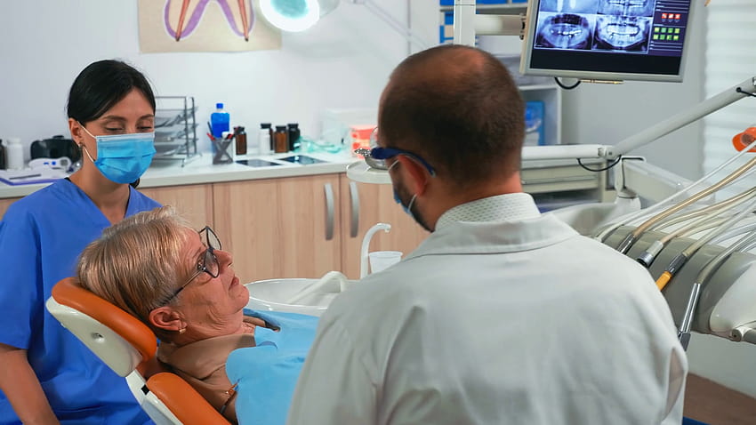 Il dentista e l'assistente lavorano con un paziente nella moderna clinica odontoiatrica. Infermiera e medico che indossano maschere di protezione che trattano i denti di una donna anziana in ufficio stomatologico sdraiata sulla sedia con la bocca aperta. Azione Sfondo HD