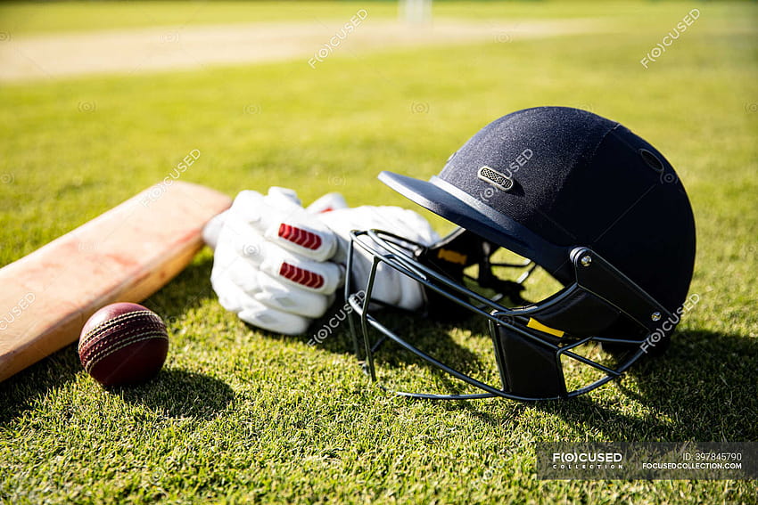 Изглед отблизо на червена топка за крикет, зелена каска за крикет, бухалка за крикет и ръкавици за крикет, лежащи на игрище за крикет в слънчев ден HD тапет