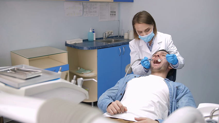Vorderansicht einer Zahnärztin mit Handschuhen und Gesichtsmaske, die hinter einem männlichen Patienten sitzt und seine Zähne mit einer Bohrmaschine in einer modernen Zahnklinik behandelt. Junger Patient wird wegen Karies behandelt Stock HD-Hintergrundbild