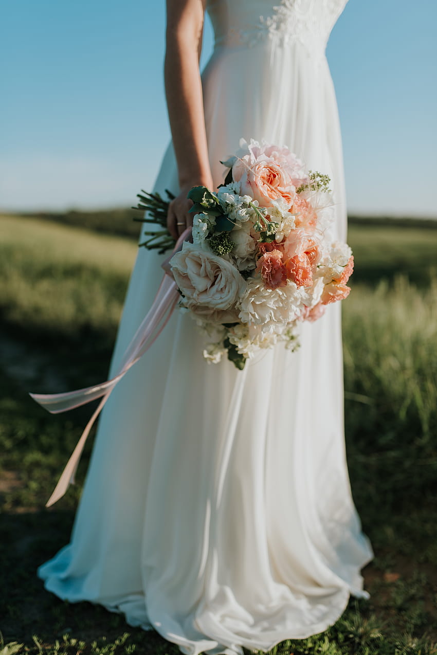 Жена, облечена в бяла булчинска рокля, държаща букет цветя, стояща на зелено поле · Дълги сватбени рокли HD тапет за телефон