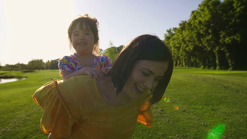 夏に屋外の緑の草の牧草地でお母さんが走っている間、お母さんの背中に乗ってダウン症の小さな幼児の女の子を笑っています。 公園で幸せな娘におんぶを与えるお母さん。 ステディカム撮影 おんぶ少年少女 高画質の壁紙
