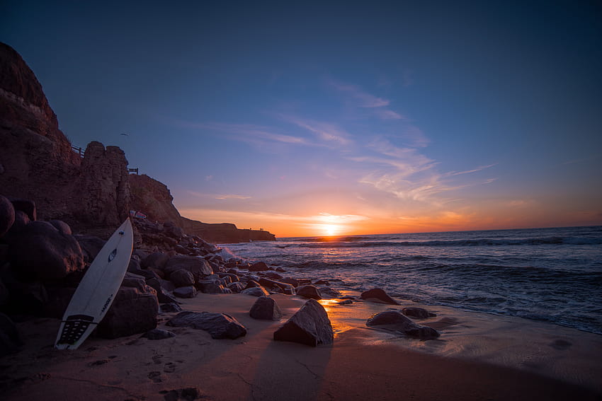 Tabla de surf blanca apoyada en roca cerca del agua al atardecer, puesta de sol de surf fondo de pantalla