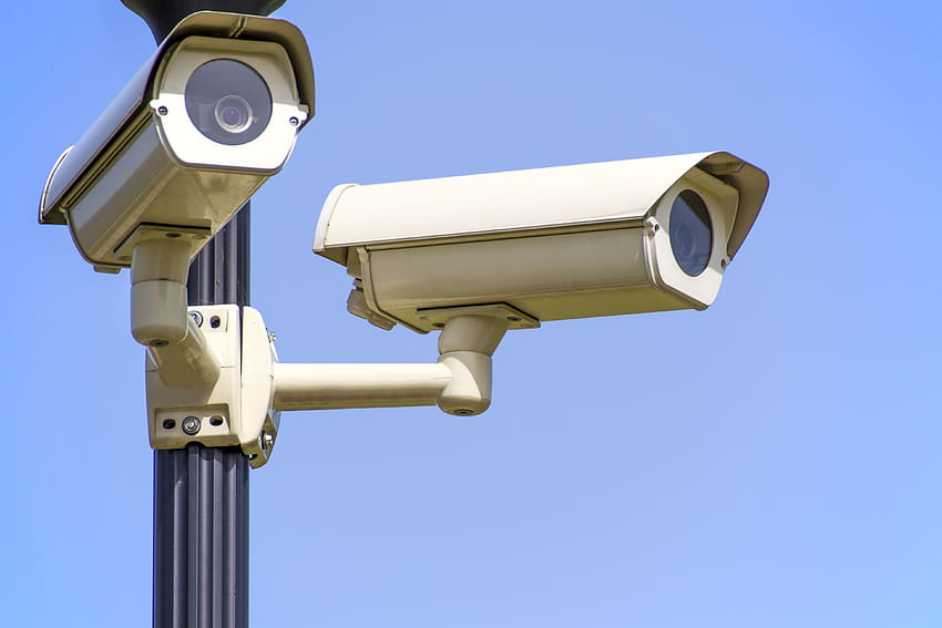 2 cámaras CCTV blancas montadas en un poste negro bajo un cielo azul despejado · Stock, vigilancia fondo de pantalla